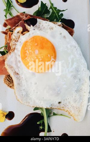 top view of fried egg with egg white and yolk, on top of a toast with bacon, arugula and dark sauce. White background, breakfast or brunch mood, Stock Photo