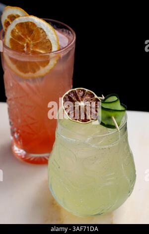 Two colorful, refreshing cocktails garnished with dried citrus slices and a cucumber ribbon, on a light marble surface against a dark background. Stock Photo