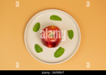 Red Apple with Mint Leaves on White Plate on creamy background, Flat view Stock Photo