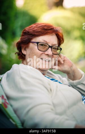 Smiling senior woman with glasses and red hair sitting in garden and enjoying her retirement and golden years. Candid portrait Stock Photo