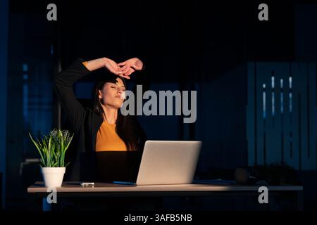 Young businesswoman or developer stretches and yawns while working late at night in a dimly lit office, showing signs of fatigue. Concept of Stock Photo