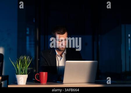 Young businessman or entrepreneur wearing glasses and a suit, working late in a dimly lit office, illuminated by laptop screen. Concept of overtime Stock Photo