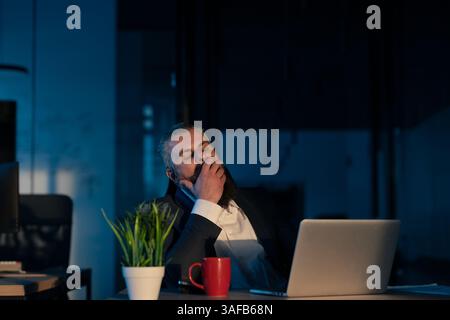 Tired entrepreneur yawning while working late at night in a dimly lit office, illuminated by laptop screen. Concept of overworking, late-night Stock Photo