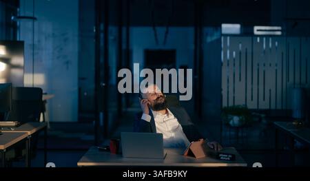 Bearded businessman in a dark office at night, wearing headphones, immersed in music while working on a laptop. Concept of work-life balance, focus Stock Photo
