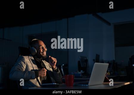 Smiling African-American man wearing headphones enjoying music at night while working on a laptop, expressing joy and relaxation. Concept of remote Stock Photo