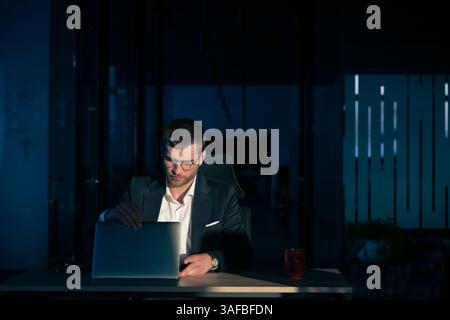 Business executive in a dark office closing his laptop, illuminated by a single light source. Concept of finishing work, exhaustion, overtime Stock Photo