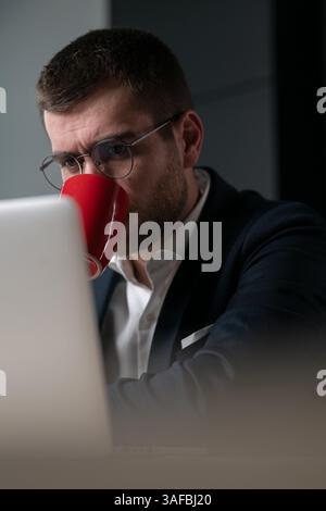 Vertical Screen: Focused man in glasses sipping coffee while working on a laptop in a modern office. Concept of concentration, productivity, and Stock Photo