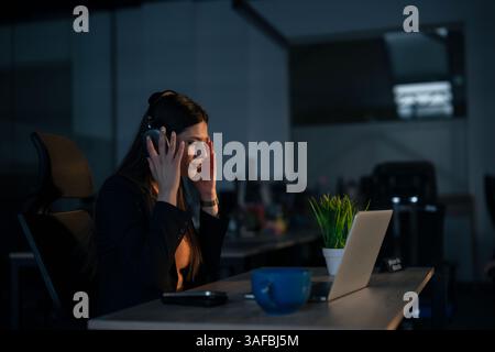 Young woman wearing headphones, enjoying music while working late in a dimly lit modern office. Concept of work-life balance, relaxation at work, and Stock Photo