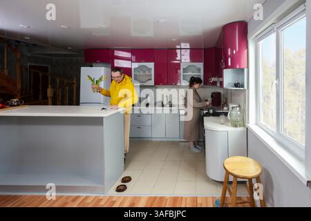 Woman cooks at stove in kitchen with pink cabinets while man looks at tulips at kitchen island in brightly lit, wide angle shot. Stock Photo