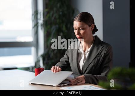 Businesswoman in a modern office opening her laptop, preparing to work with a mix of focus and mild anxiety. Concept of professional challenges Stock Photo