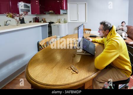 Man in yellow hoodie works on laptop at table while woman relaxes on couch nearby, comfortable home setting with kitchen in background. Stock Photo
