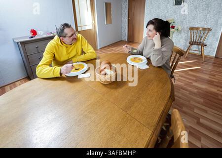 Man in yellow sits across from woman as they share food at wooden table, plates of soup visible in casual, comfortable room with natural light. Stock Photo
