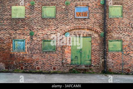 Front elevation brick wall of now disused historical warehouse with green painted boarded windows and one glass window with reflections of sky, tiles. Stock Photo