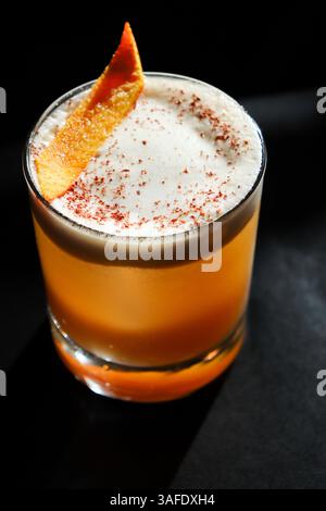 Close-up of a classic whiskey sour cocktail topped with frothy egg white foam and garnished with a fresh orange peel, served in a clear glass Stock Photo