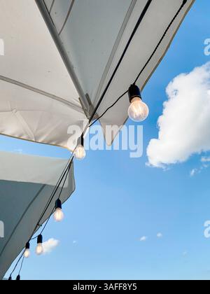 Looking up at the outdoor cafe umbrella, blue sky with one cloud in the background. White caffee umbrella with few glowing edison light bulbs. Stock Photo