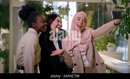 Three diverse multiracial business ladies in office happy girls smiling posing mobile phone camera students Asian Muslim African businesswomen Stock Photo
