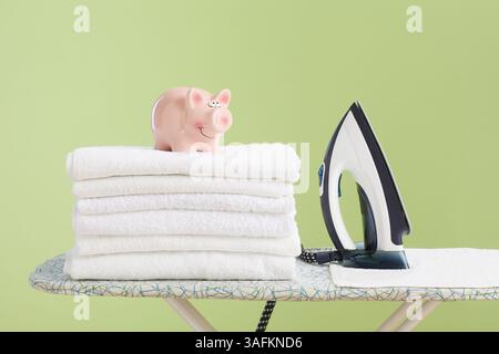 A pink piggy bank and standing iron placed on a stack of folded white towels on an ironing board against a green background, symbolizing domestic savi Stock Photo