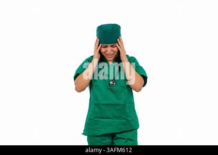 Young stressed latin surgeon wearing green uniform and stethoscope suffering from headache touching temples with closed eyes on white background, expr Stock Photo