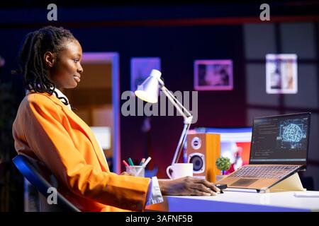 Smiling woman using laptop in apartment, updating artificial intelligence machine learning algorithms, writing code. Upbeat teleworking IT expert working from home uses programming to upgrade AI Stock Photo