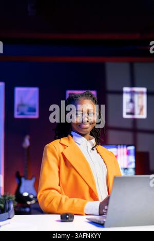 Portrait of jolly woman sitting on chair at home office desk in neon illuminated apartment late at night. Cheerful african american person in RGB lit living room enjoying leisure time Stock Photo