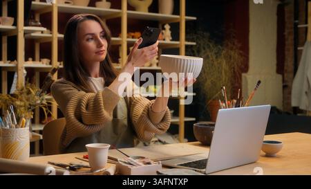 Caucasian woman girl female potter sculptor artist blogger using mobile phone handmade bowl photo in workshop crafting hobby small business owner sale Stock Photo