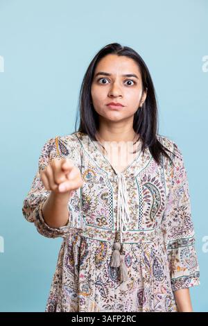 Indian serious woman pointing her index finger at the camera and looking forward, choosing someone and acting calm against blue background. Young person satisfied making a choice. Stock Photo