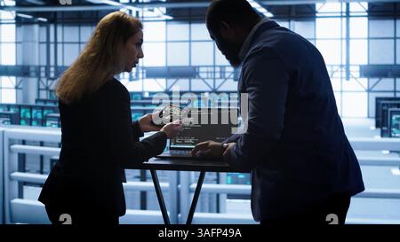 Team of developers doing maintenance checkup using laptop to prolong data center hardware life span. IT support staff members in server hub monitor server infrastructure using notebook, camera A Stock Photo