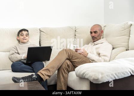 Father using smartphone and son using laptop, both sitting on comfortable sofa at home, enjoying technology and family time Stock Photo