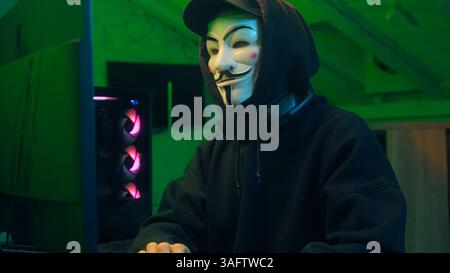 Lviv, Ukraine - June 03, 2024: Keyboard computer hacking man mask anonymous typing desktop pc looking at camera steal monitor database coding virus Stock Photo
