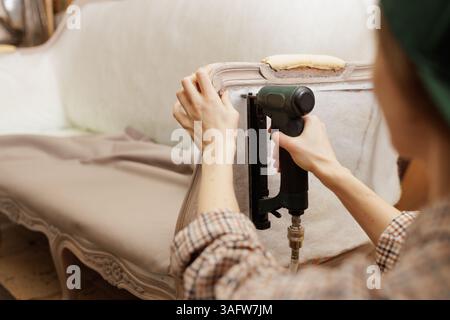 Close-up of woman upholstering vintage sofa with pneumatic stapler, focusing on craftsmanship and detail. Stock Photo