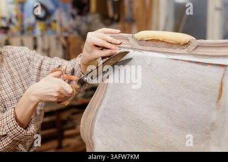 Close-up of hands cutting upholstery fabric with large scissors while restoring vintage furniture in a workshop. Stock Photo
