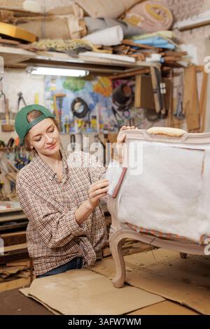 Young female upholsterer sanding vintage chair in workshop, restoring furniture with precision and creativity. Stock Photo