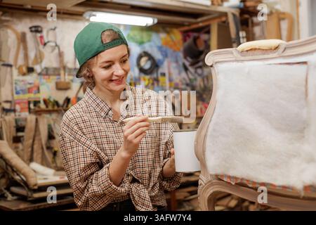 Smiling female artisan applying paint to vintage chair frame during upholstery restoration in workshop. Stock Photo