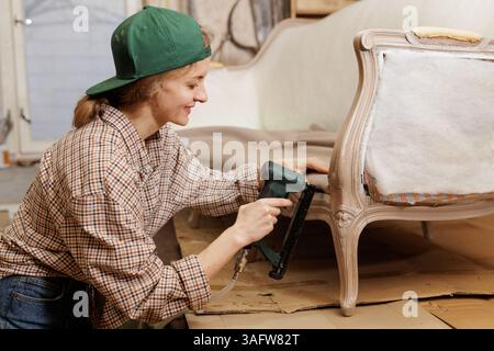 Smiling female upholsterer using a pneumatic staple gun while restoring a vintage armchair in a workshop. Stock Photo