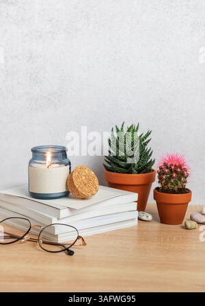 Cozy home interior decor with burning scented candle, stack of books, eyeglasses, and potted plants on wooden table Stock Photo