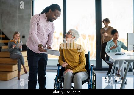Disabled woman in wheelchair looking at computer, discussing startup project with interracial team Stock Photo