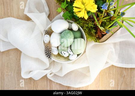 Stylish decorated easter eggs in a white bowl and spring primroses in a pot on linen cloth on a light wooden table. Top view. Happy Easter concept Stock Photo