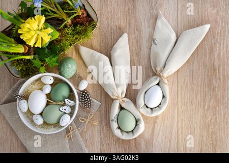 Easter eggs in a white bowl on linen cloth and spring primroses in a pot. Easter eggs wrapped in a napkins folded like bunny ears. Happy Easter concep Stock Photo