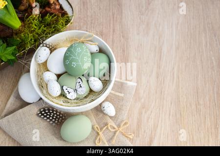 Stylish decorated easter eggs in a white bowl on linen cloth on a light wooden table. Top view. With space for your text Stock Photo