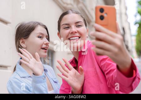 Two caucasian women waving at smartphone camera while taking selfie outdoors Stock Photo