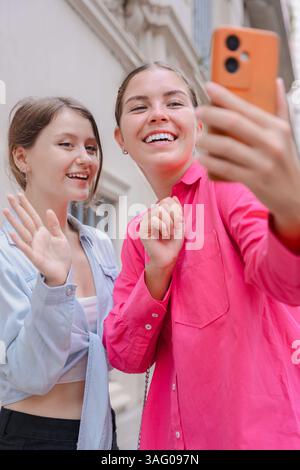 Two caucasian women waving at smartphone camera while taking selfie outdoors. Stock Photo