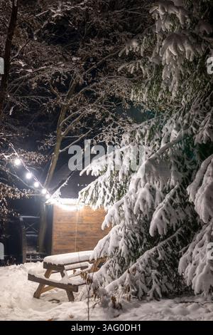 A snow-covered wooden picnic table and benches stand in a forest at night, illuminated by string lights hanging in the background Stock Photo