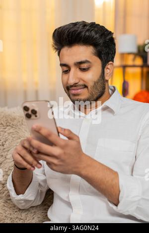 Indian young man at home sitting on sofa focused on smartphone typing text, browsing news, reacting to notifications. Arabian guy using mobile phone for social media, work, texting, digital lifestyle Stock Photo