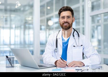 A friendly doctor wearing a stethoscope smiles while sitting at his desk in an office setting, ready for consultations. Stock Photo