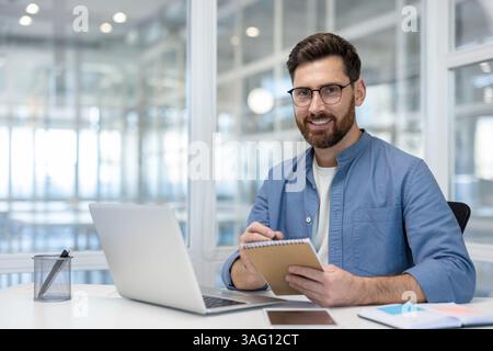 A smiling man with a beard and glasses working in a modern office setting, focused on his notepad. He is surrounded by a laptop and other office supplies. Stock Photo