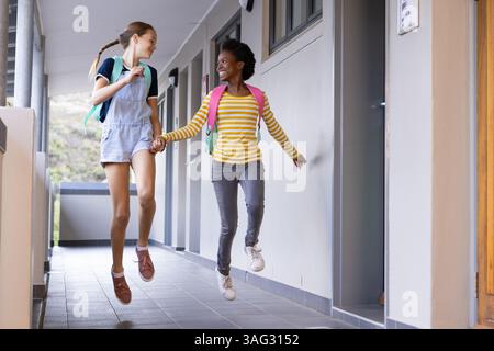Jumping and holding hands, two diverse schoolgirls with backpacks walking in hallway Stock Photo