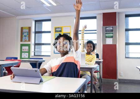 In school, diverse students raising hands while using tablet in classroom Stock Photo