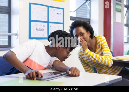 In school, Whispering secrets, diverse students in classroom sitting at desk with laptop Stock Photo