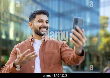 A smiling man holds up his phone, possibly video-calling or taking a selfie, outside a modern building. Stock Photo