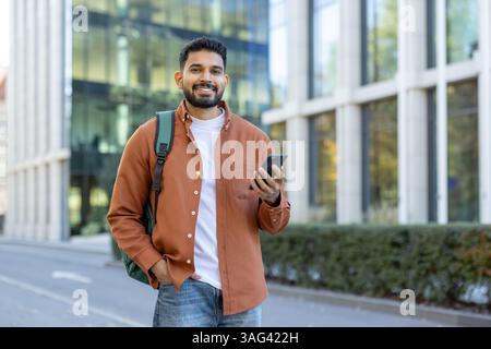A smiling man with a backpack holds his smartphone outside a modern building, enjoying the sunny day in the city. Stock Photo